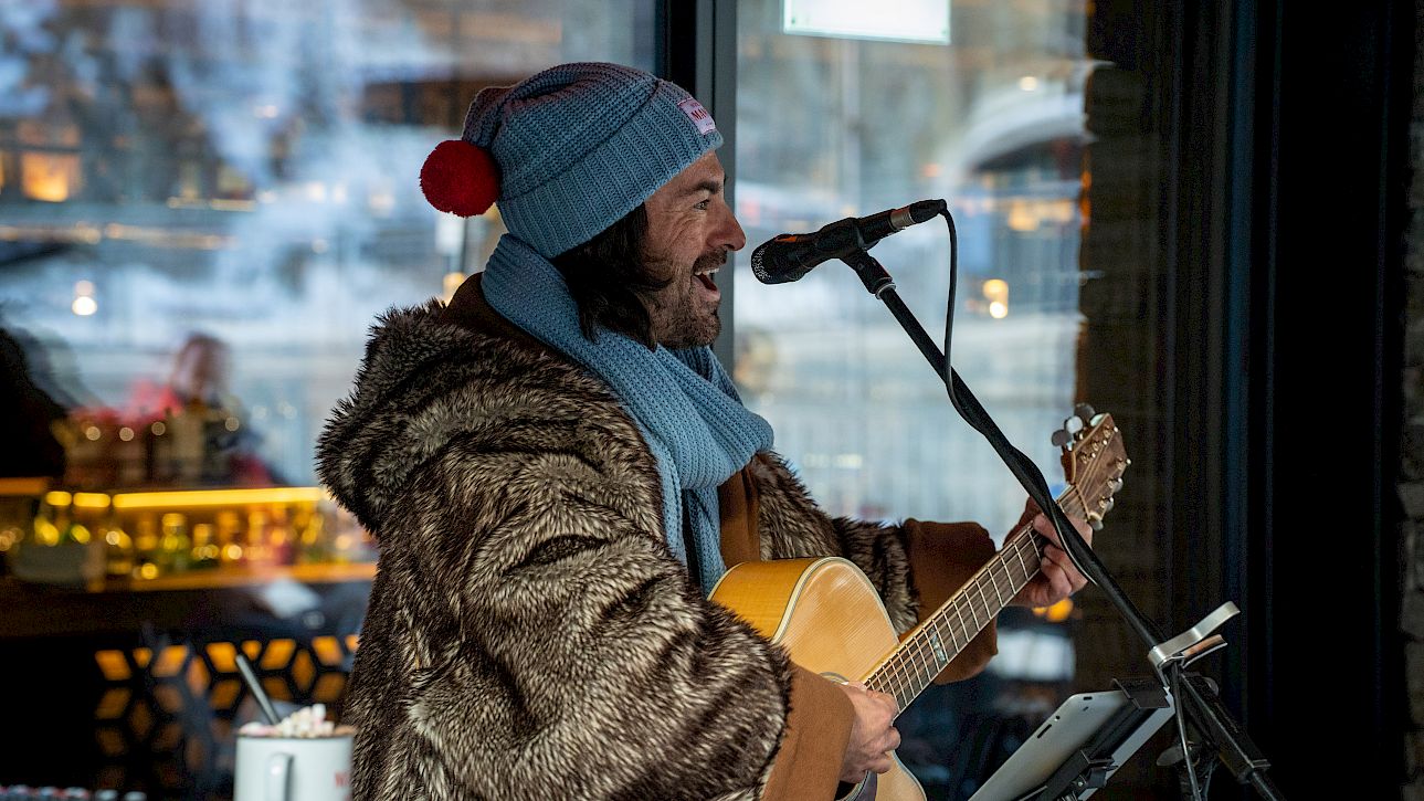 Musician in winter clothes playing guitar and singing in a cozy café with a view of snow-covered trees.
