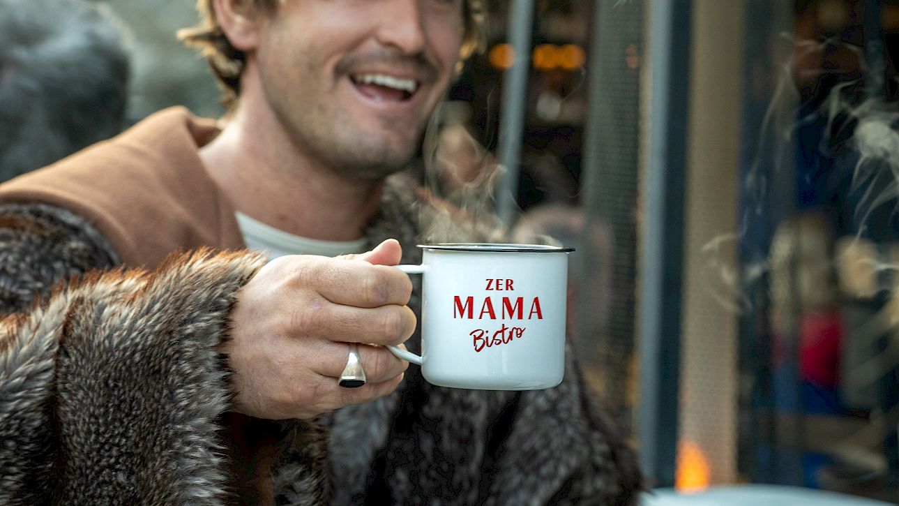 A man in a fur jacket and cap holds a mug with "Der Mama Bär" written on it.