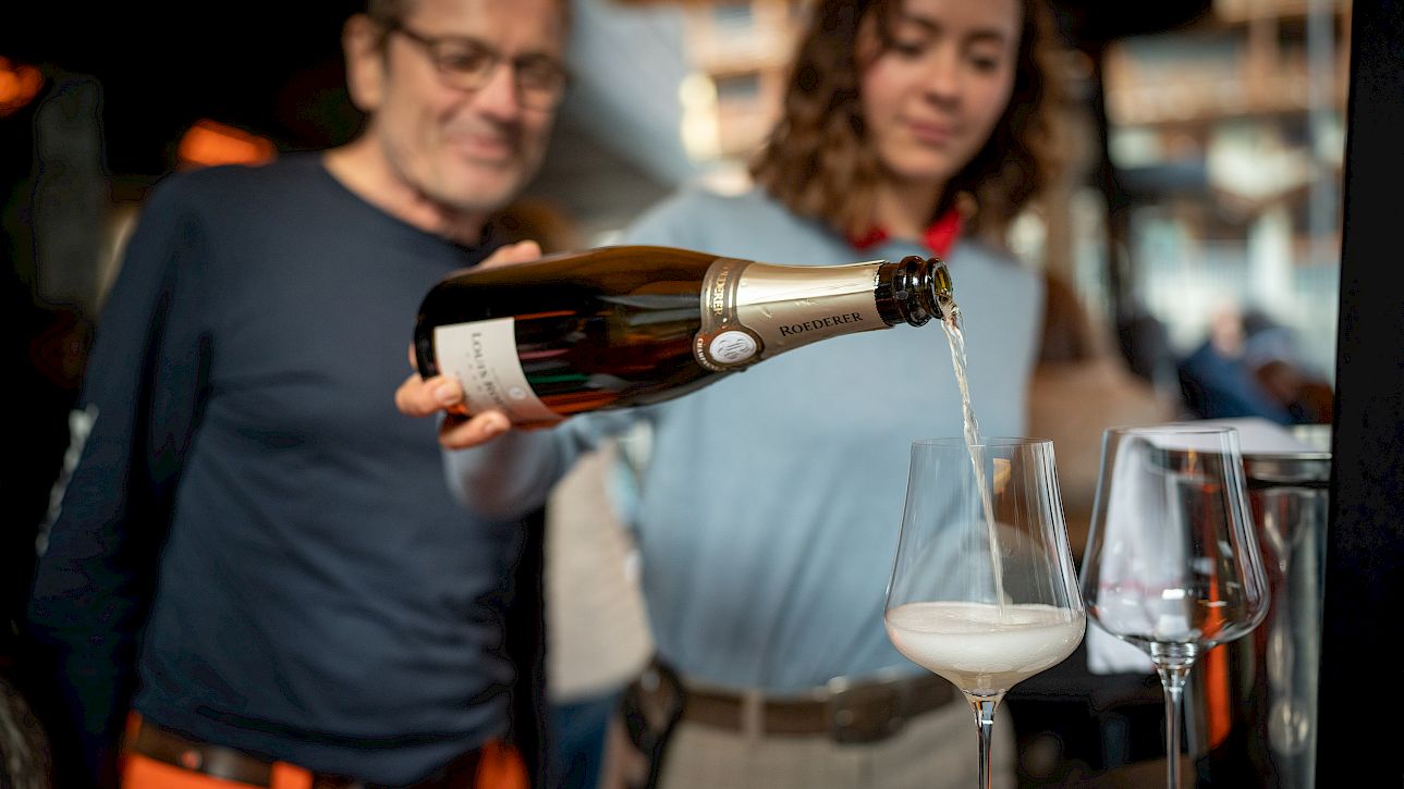 A woman pours champagne into two glasses in an elegant setting, while a man watches with interest.