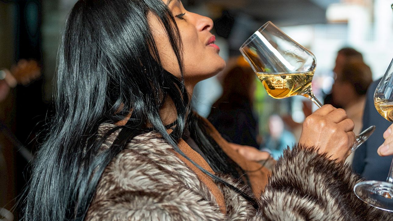 Woman in fur coat raises a wine glass at an outdoor gathering, appearing to savor the moment.