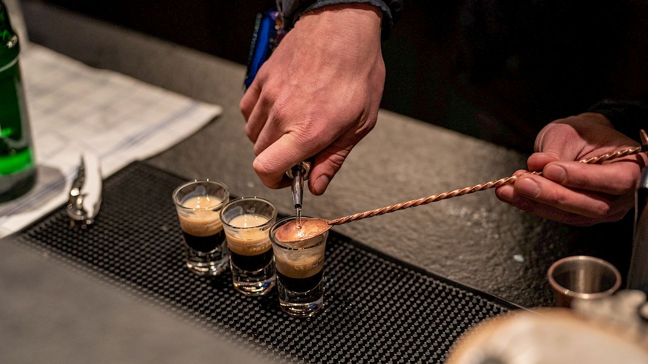 A bartender prepares three layered espresso shots on a counter using a long spoon.
