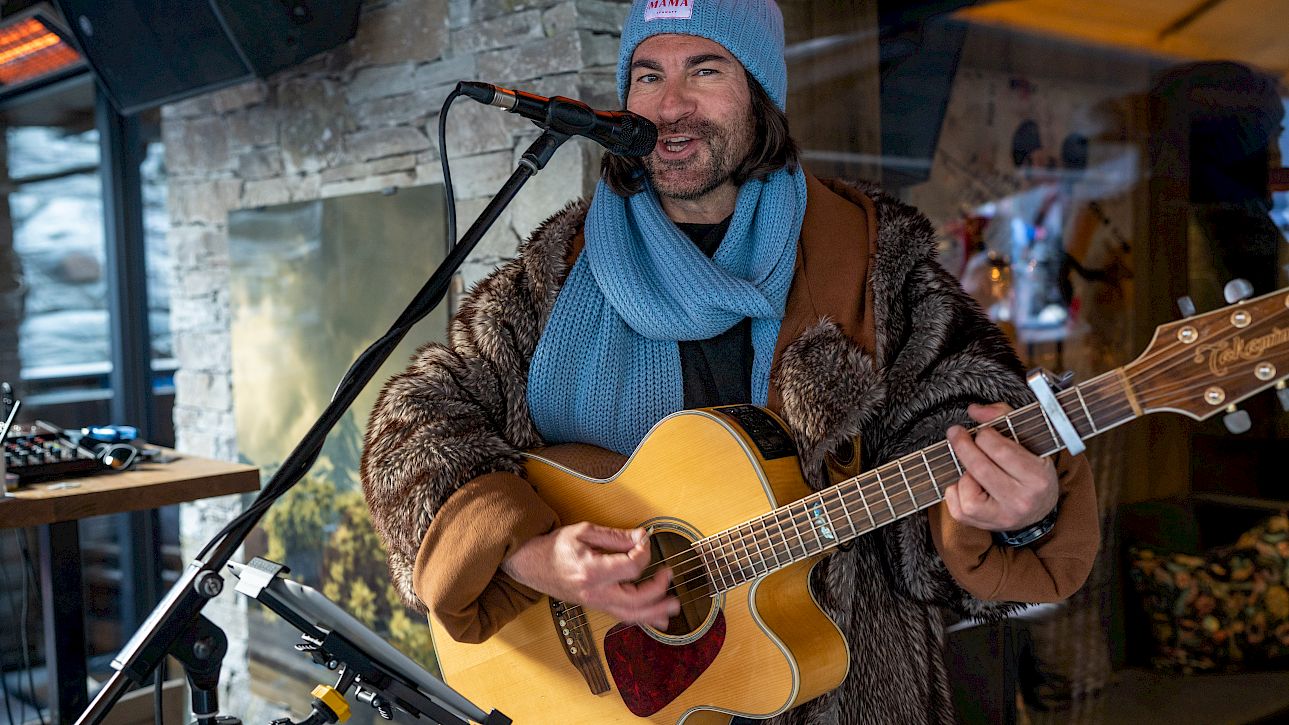 Man in winter clothes plays guitar and sings into a microphone, with a stone wall in the background.
