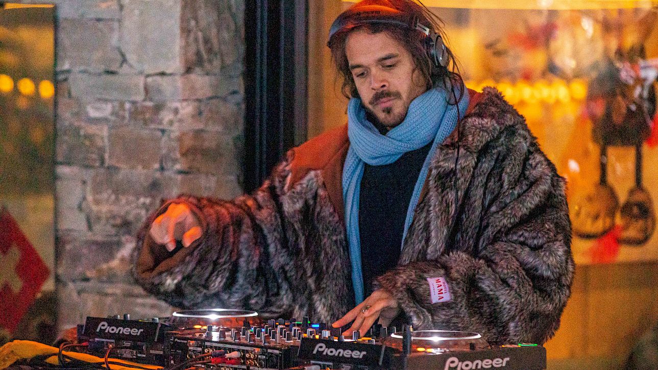 DJ performing outdoors, wearing a fur coat and blue scarf while mixing music.