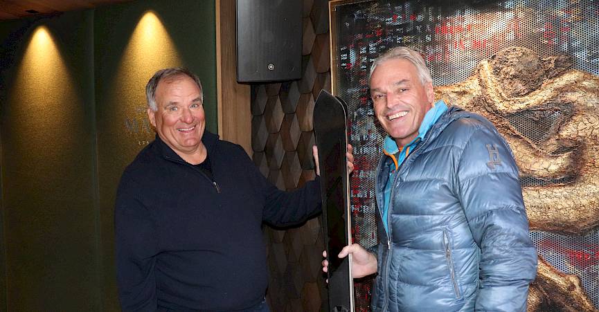 Two men indoors posing with a ski, smiling and casually dressed.
