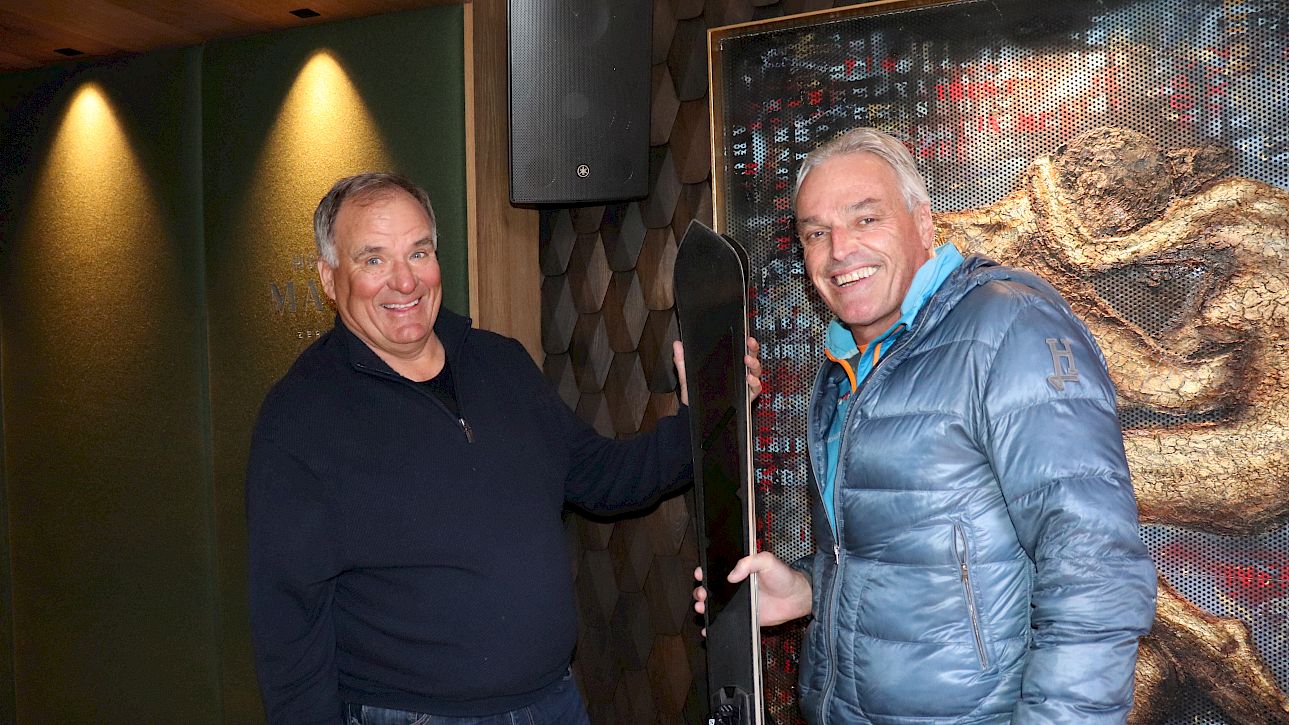Deux hommes souriants tiennent ensemble un snowboard devant un fond artistique.