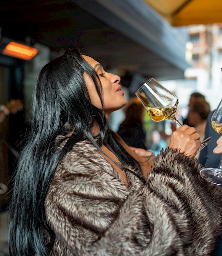 Eine Frau in einem Pelzmantel genießt ein Glas Weißwein bei einer Veranstaltung im Freien.