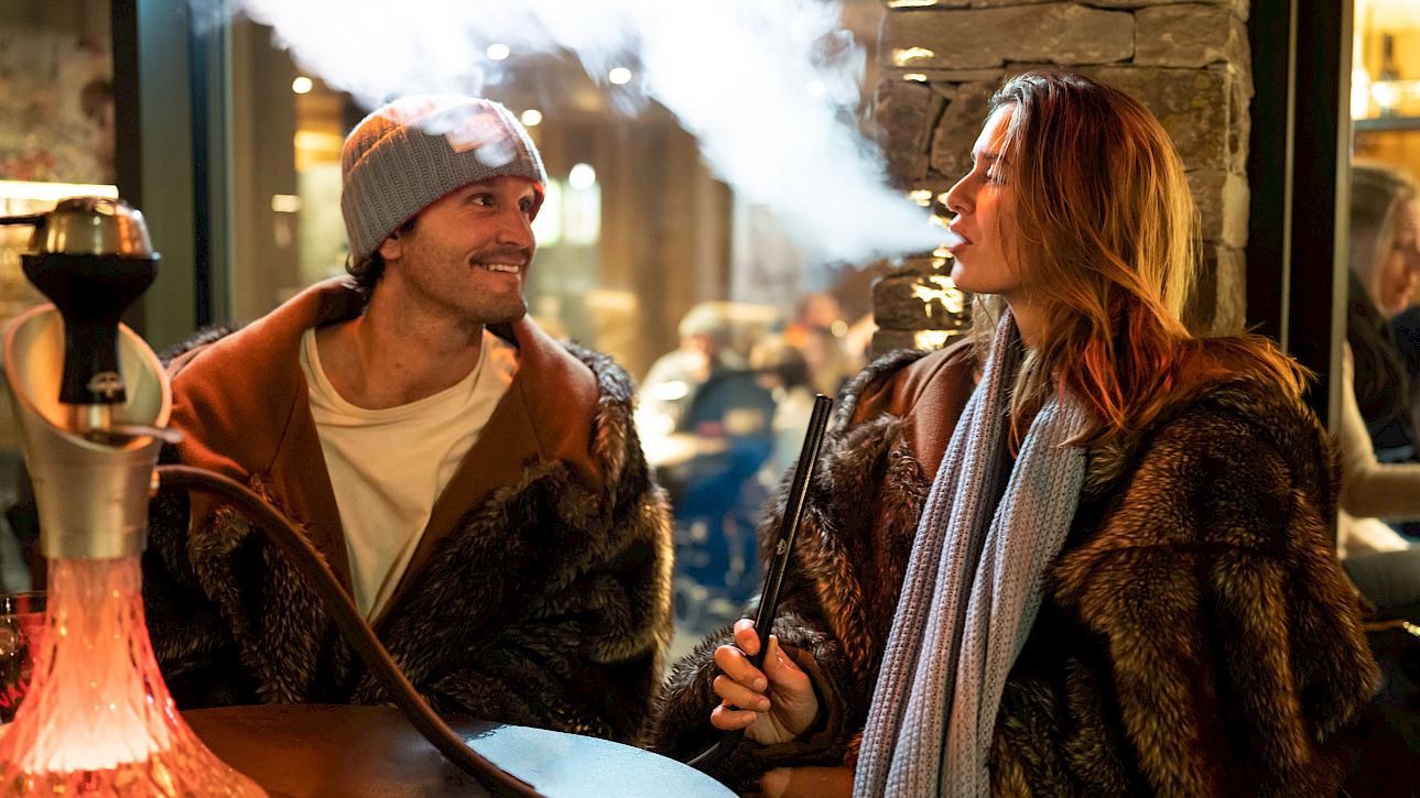 Couple en tenue d'hiver fume la chicha dans un bar chaleureux. La femme souffle de la fumée en l'air.
