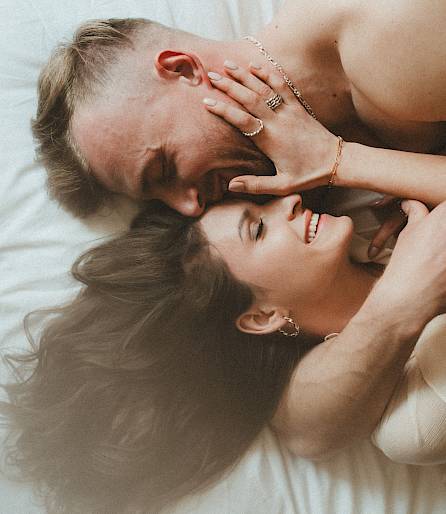 A smiling couple lies close together on the bed, enjoying an intimate moment.