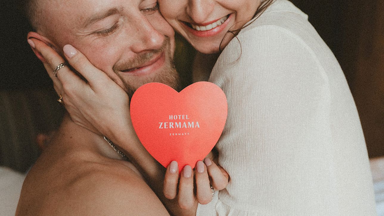 Un couple s'embrasse tendrement, tenant un cœur rouge avec "Hotel Zermama" écrit dessus.