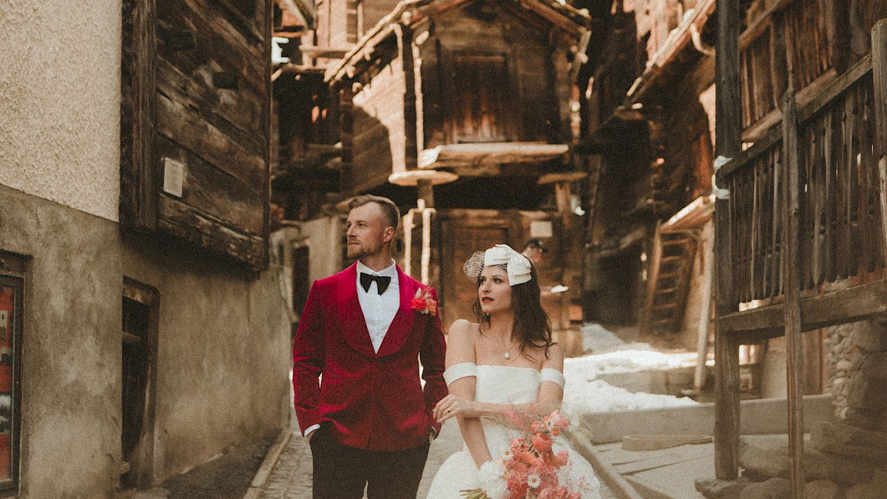 Un couple de mariés se promène dans une ruelle pittoresque en montagne, entouré de vieilles maisons en bois.