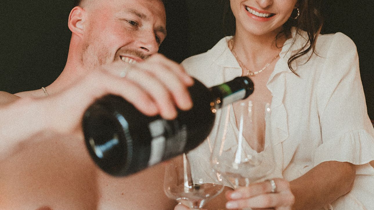 Un couple au lit, l'homme sert du champagne dans deux verres pendant que la femme regarde en souriant.