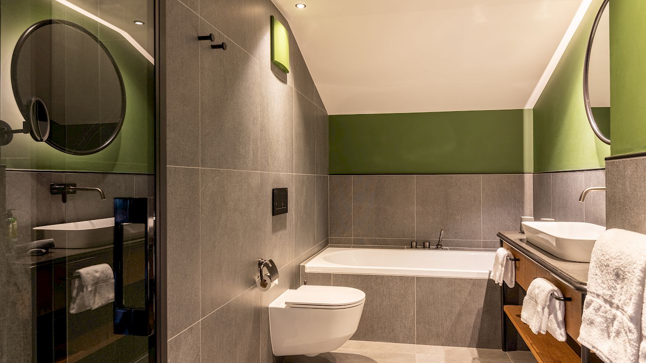 Modernes Badezimmer mit grauen Fliesen, grünen Akzenten, runder Spiegel, Badewanne und Holzwaschtisch.