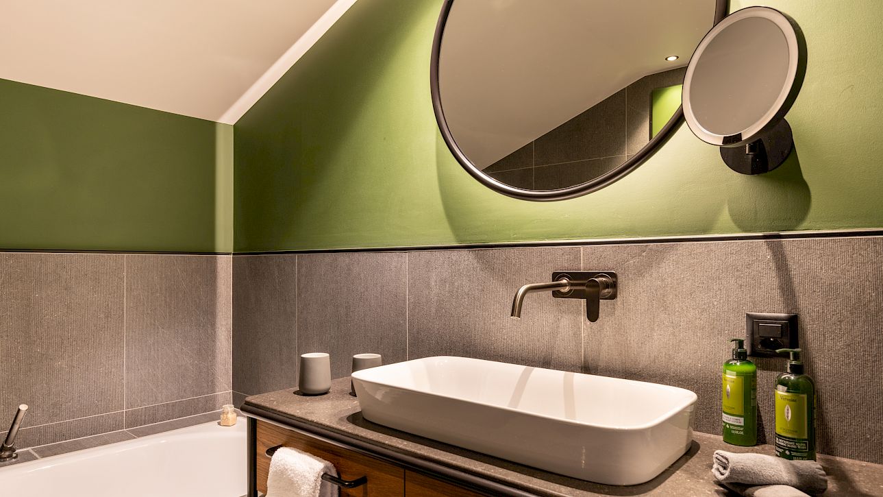 Modernes Badezimmer mit grüner Wand, ovalem Spiegel, Waschschale und Badewanne, elegante und schlichte Einrichtung.