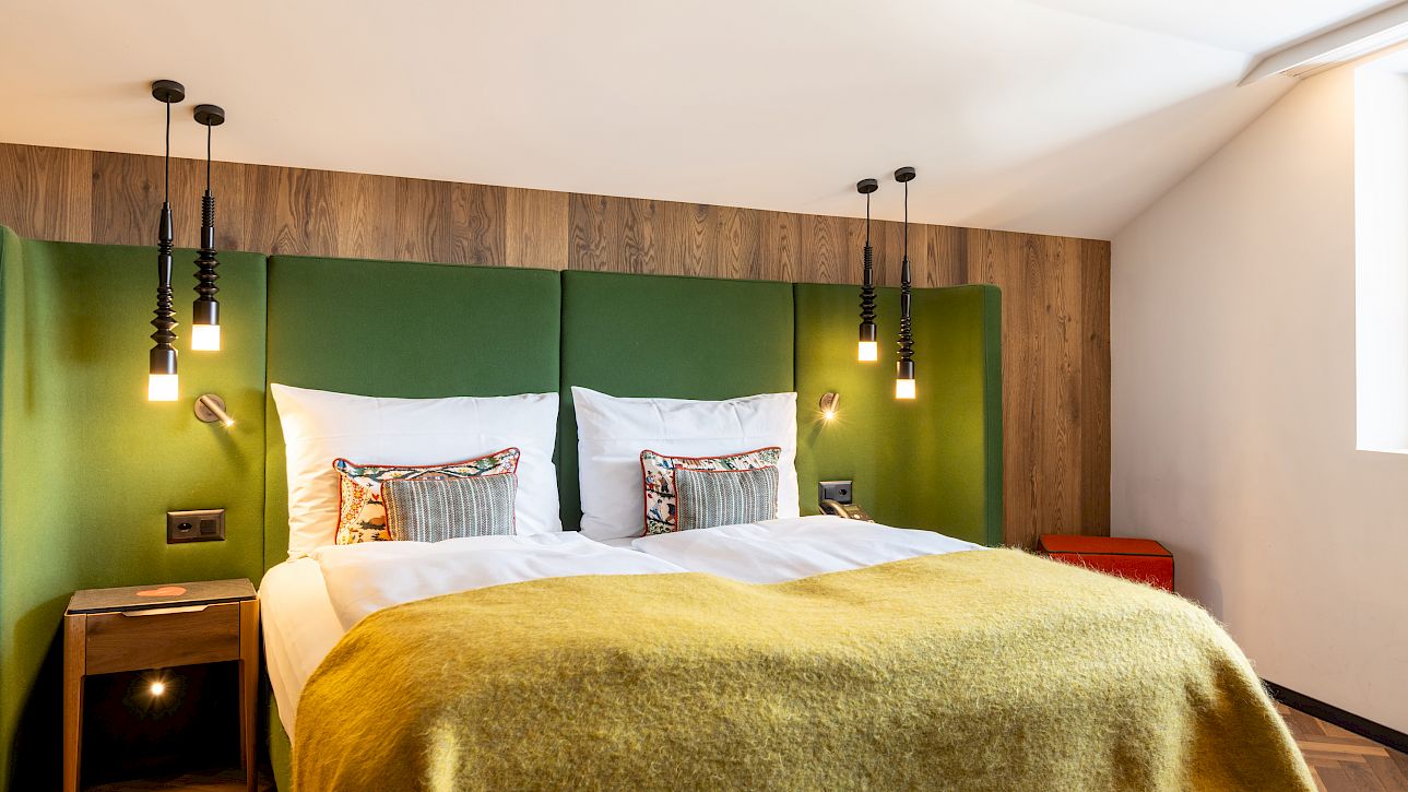 Gemütliches Hotelzimmer mit Doppelbett, grüner Kopfleiste, hängenden Lichtern und Holztönen.