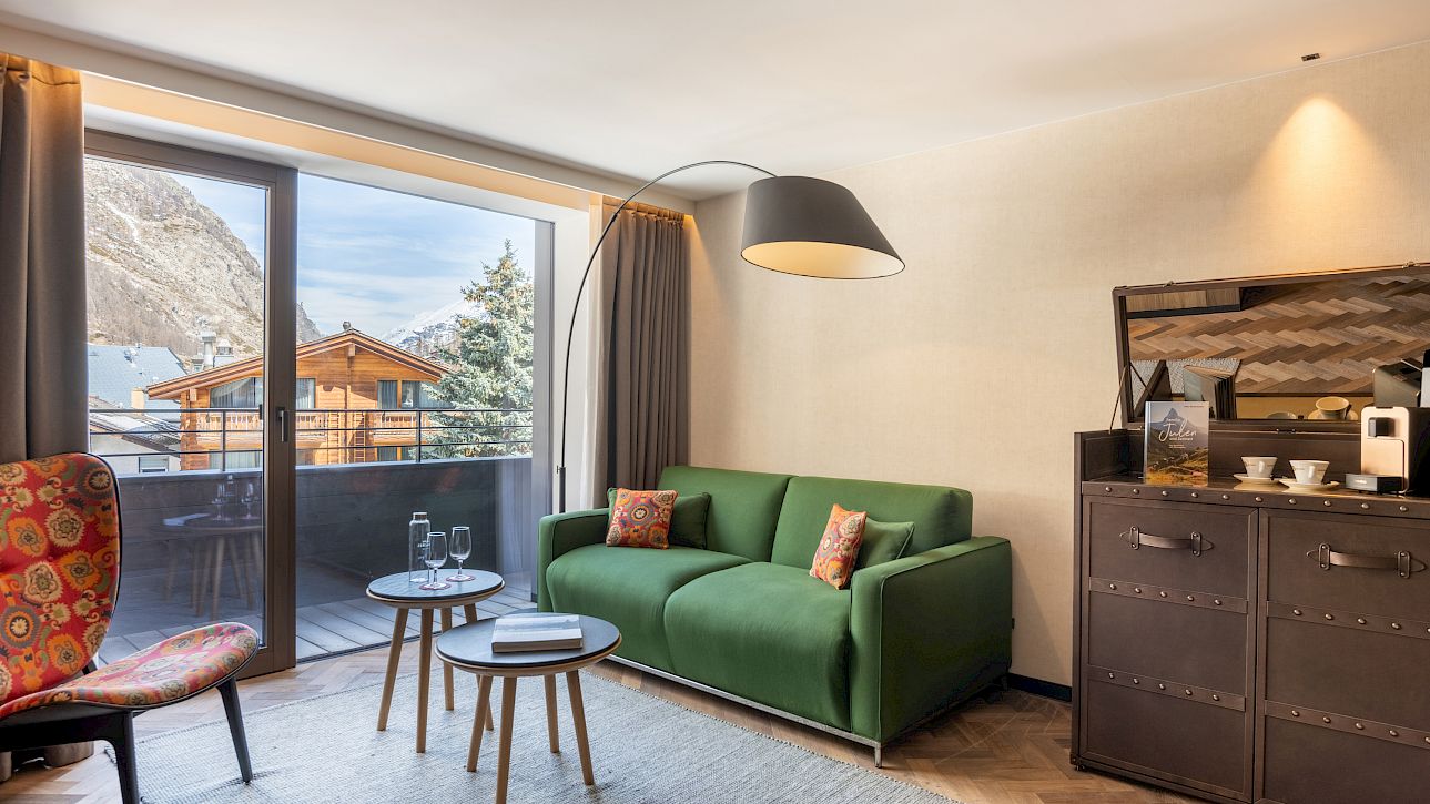 Gemütliches Wohnzimmer mit grüner Couch, Balkonblick auf Berge, stilvolles Dekor, Kaffeemaschine rechts.