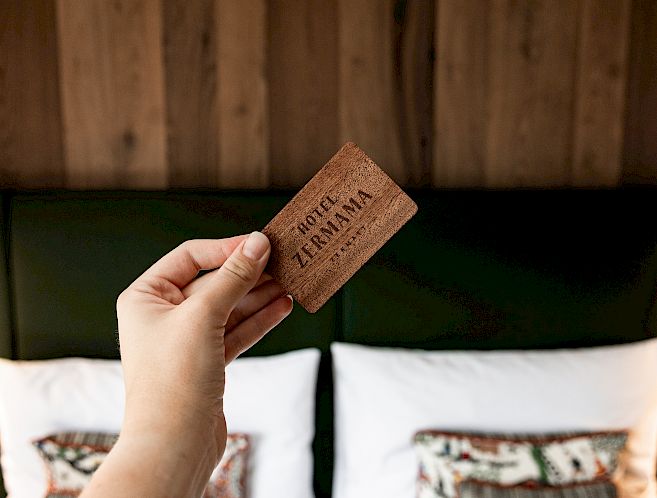 Eine Hand hält eine Zimmerkarte vor einem Doppelbett mit dekorativen Kissen und Holzwand im Hotelzimmer.
