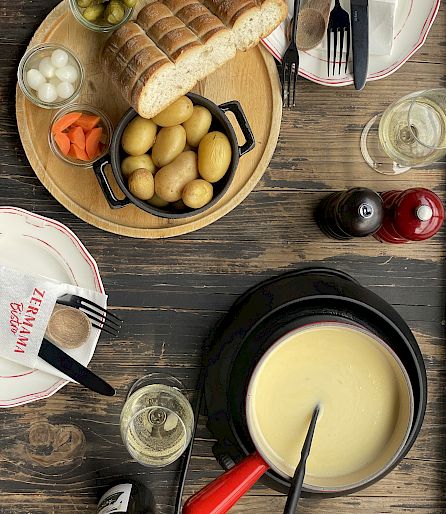 Fondue-Set auf Holztisch mit Brot, Kartoffeln, Gemüse und Wein. Zwei Gedecke mit Besteck sind sichtbar.