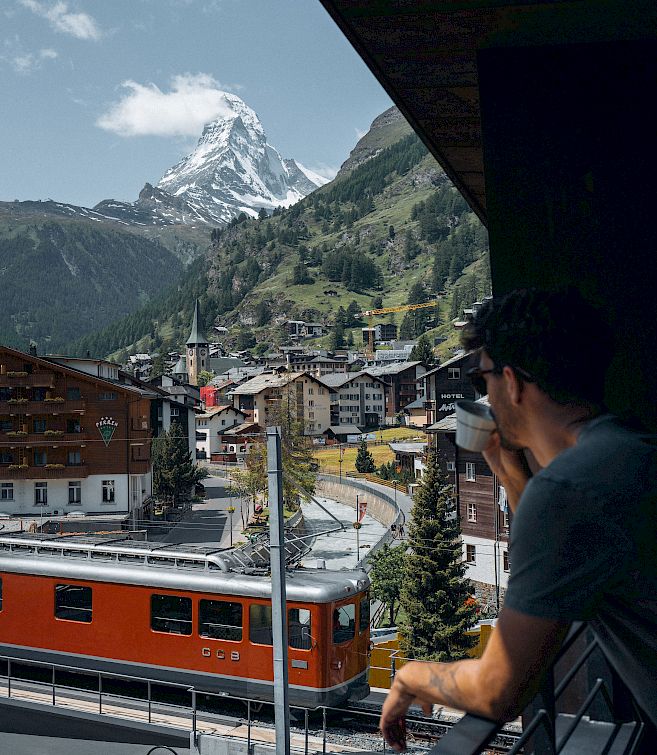 Mann betrachtet den Matterhorn-Gipfel von einem Balkon aus, während ein roter Zug durch das Dorf fährt.