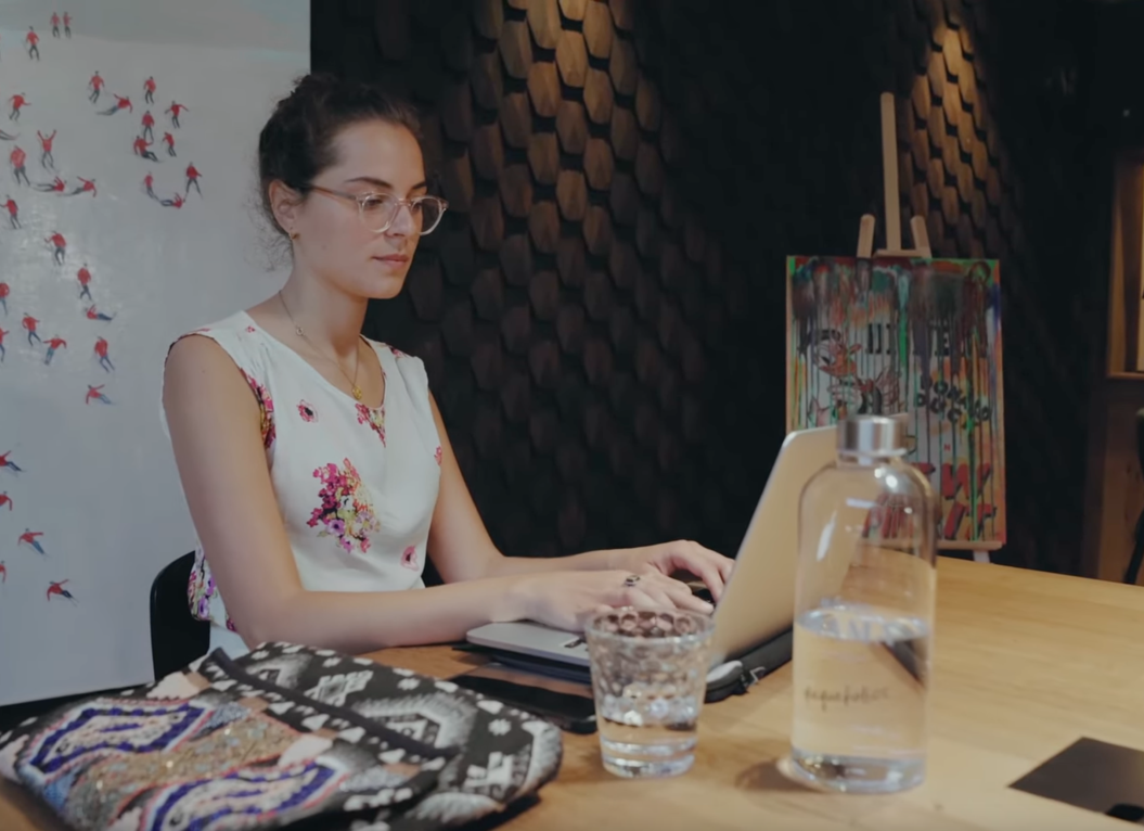 Frau mit Brille arbeitet am Laptop, Kunstwerke und Wasserflasche auf einem Holztisch im modernen Raum.