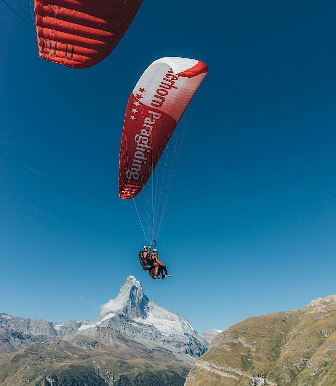 Personen beim Gleitschirmfliegen vor dem Matterhorn, blauer Himmel und Berglandschaft im Hintergrund.