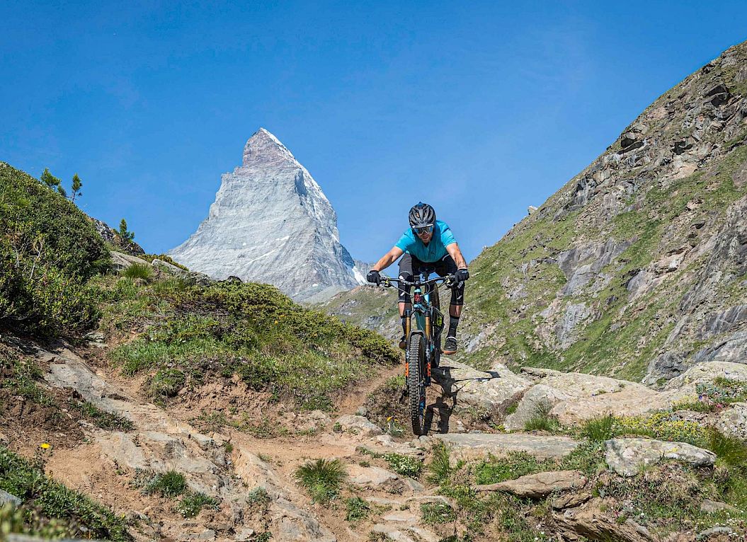Radfahrer fährt einen steinigen Bergpfad hinab, mit Blick auf das Matterhorn unter klarem blauem Himmel.