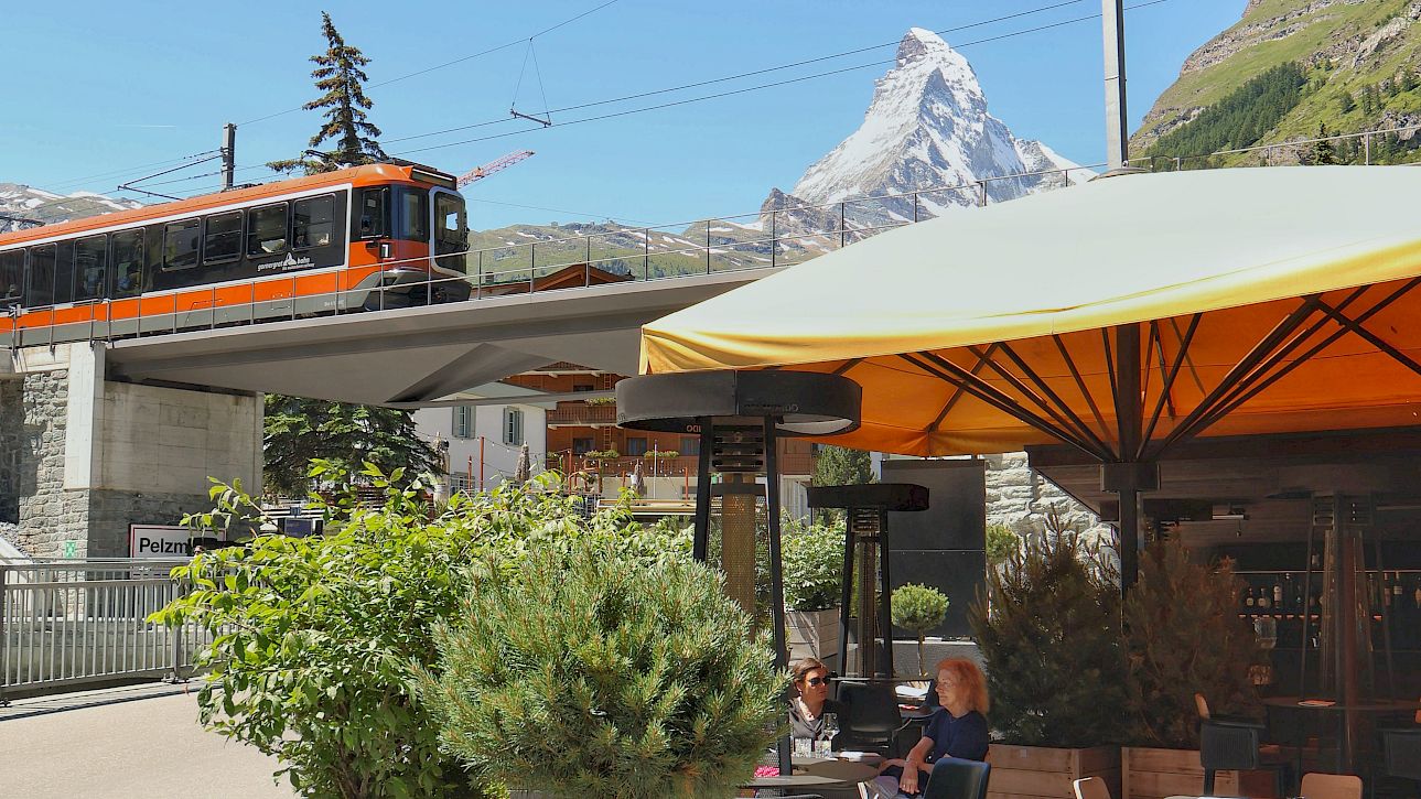 Café-Terrasse mit Blick auf Zug und schneebedeckten Berg bei klarem Himmel. Pflanzen im Vordergrund.