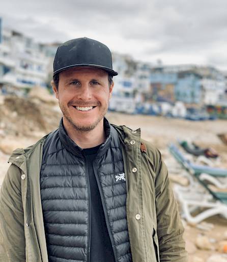 Person am Strand mit beiger Jacke und schwarzer Mütze, im Hintergrund verschwommene Gebäude.