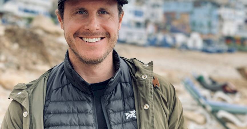 Personne sur une plage avec une veste beige et une casquette noire, bâtiments flous en arrière-plan.