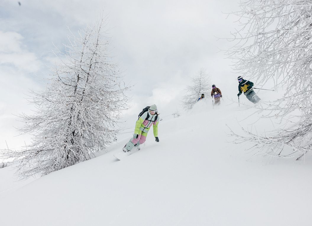 Snowboarder fahren eine verschneite Piste hinunter, flankiert von schneebedeckten Bäumen.