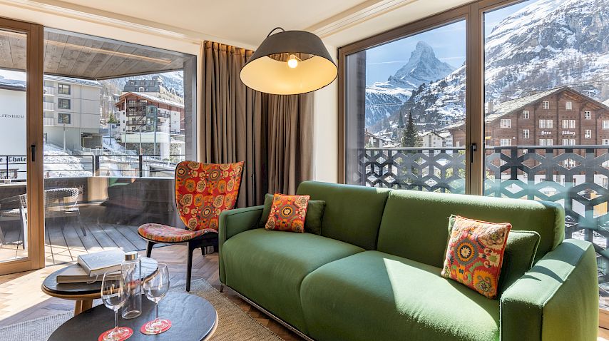 Wohnzimmer mit grünem Sofa, Balkon und Matterhornblick. Moderne Einrichtung, helle Fenster.
