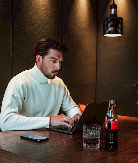 Ein Mann im weißen Pullover arbeitet an einem Laptop. Auf dem Tisch stehen Getränke und Blumendekoration.