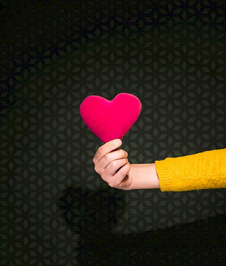 Ein Arm in gelbem Pullover hält ein rotes Stoffherz vor schwarzem Hintergrund mit Muster.