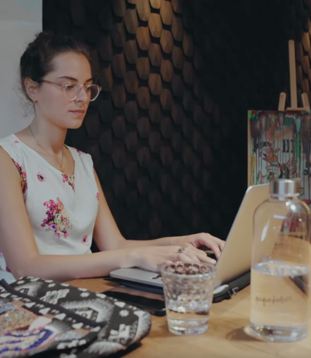 Jeune femme avec lunettes assise à une table, travaillant intensément sur un ordinateur portable dans une pièce élégante.