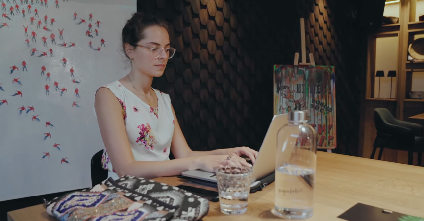 Young woman with glasses sits at a table, working intently on a laptop in a stylish room.