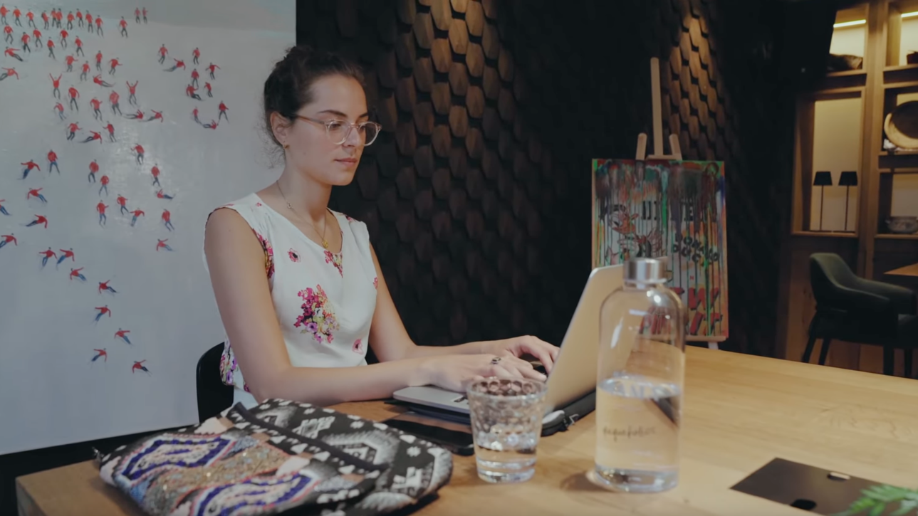 Femme avec des lunettes travaillant sur un ordinateur portable dans une pièce moderne, avec un verre d'eau à côté.
