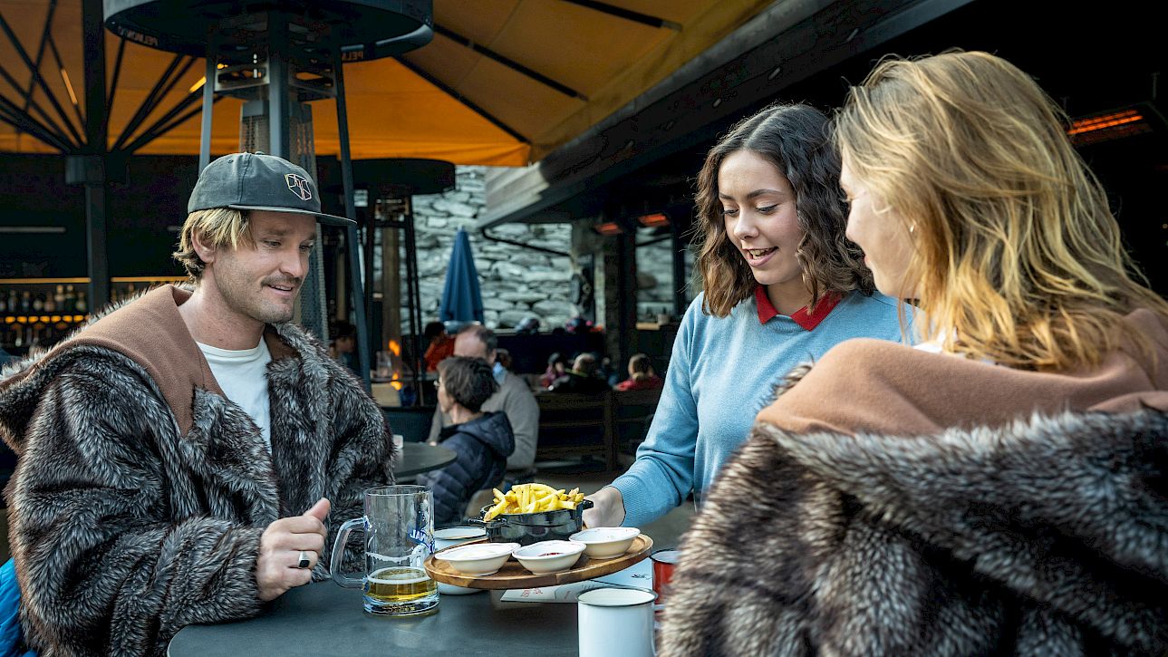 Three people in fur coats enjoy snacks and drinks at an outdoor table under yellow umbrellas.