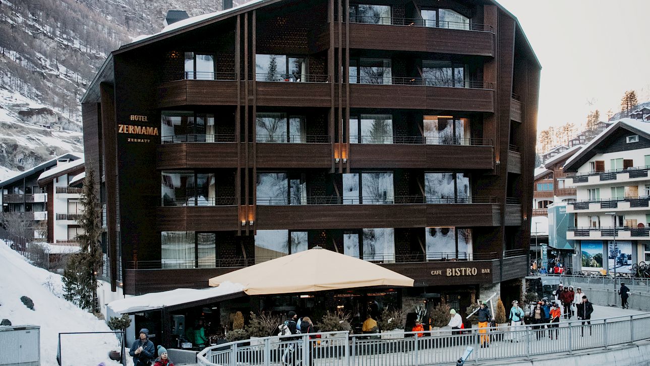 Wooden building in a snowy mountain town, people walking, some in ski gear.