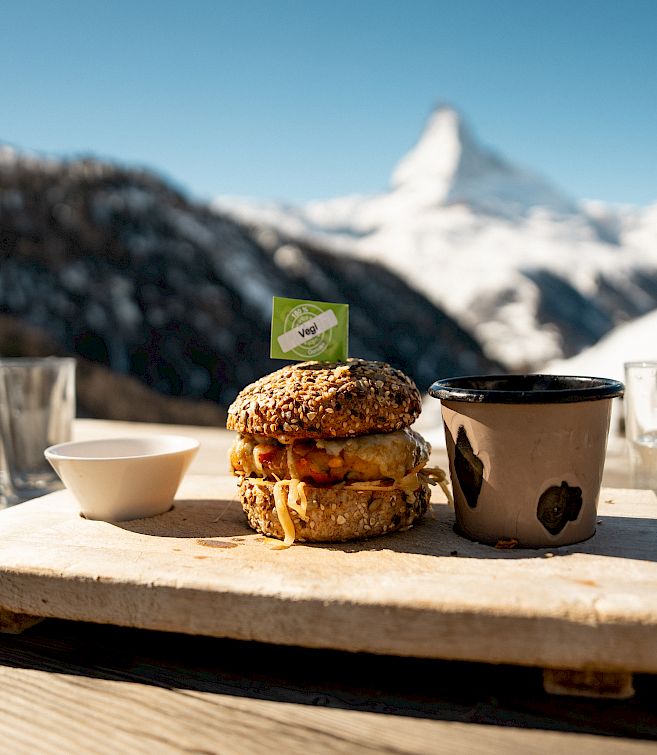 Burger on wooden board with cup and glass, snowy mountain backdrop, sunny view.