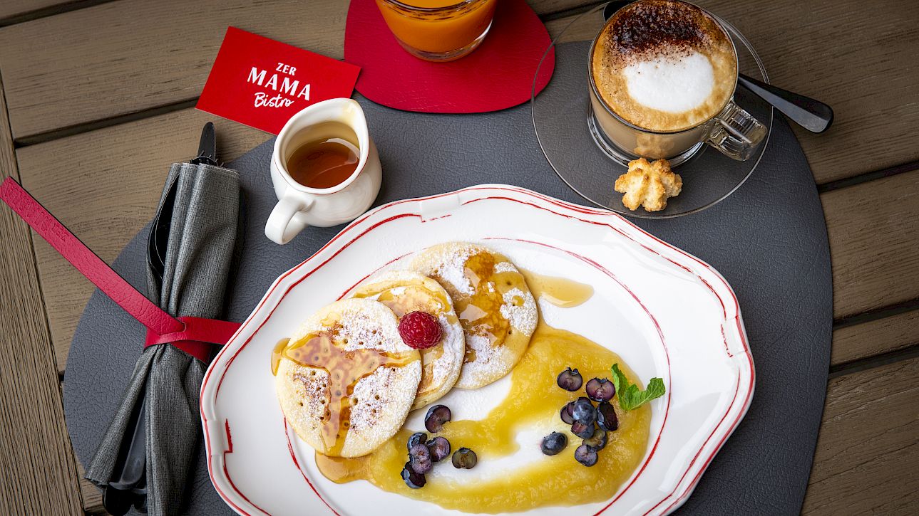 Pancakes with berries and syrup on a plate, next to coffee and orange juice.
