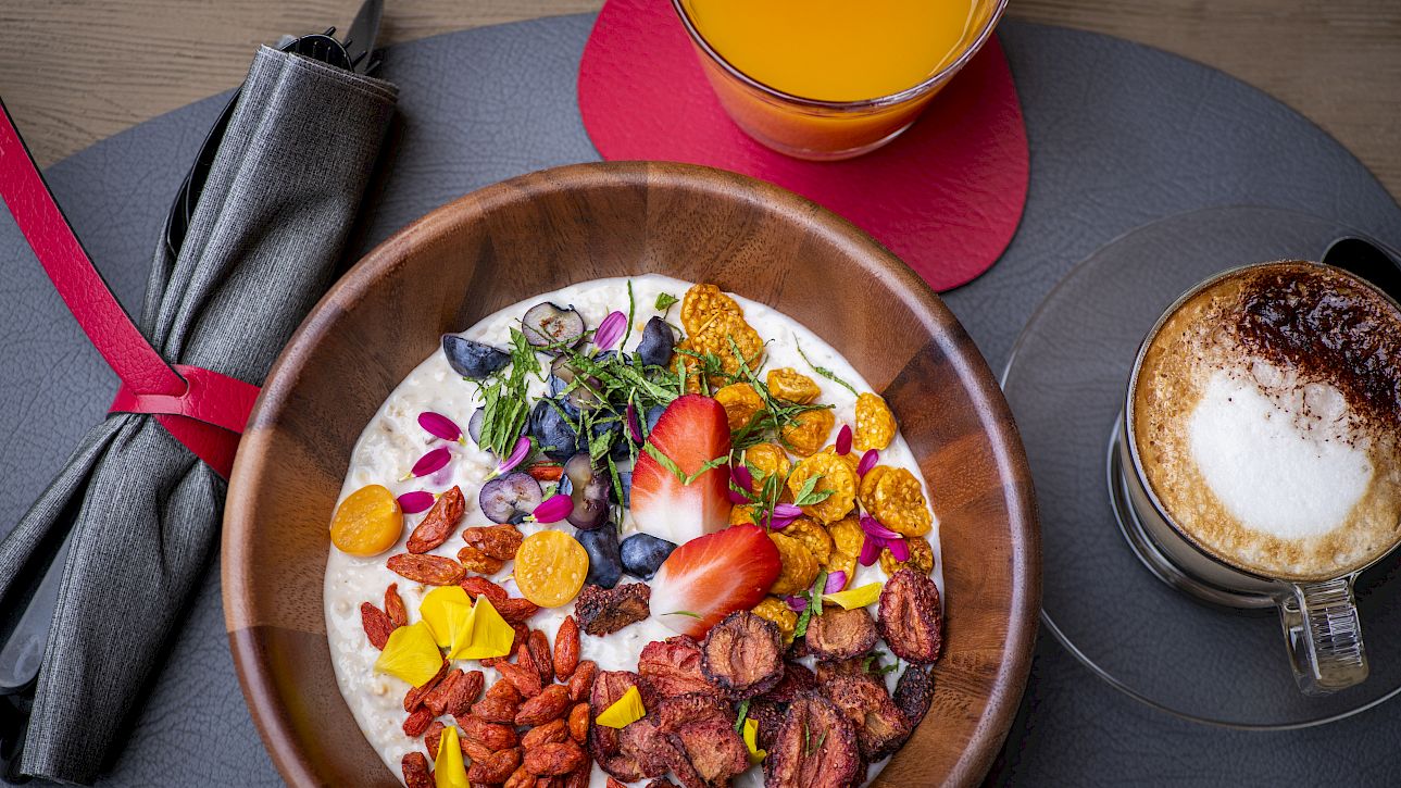 Petit-déjeuner avec bol de muesli aux fruits, cappuccino et jus d'orange sur set de table gris.