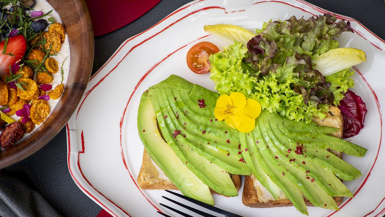 Avocado toast with salad and flowers, next to a bowl of cereal with berries, arranged on a table.