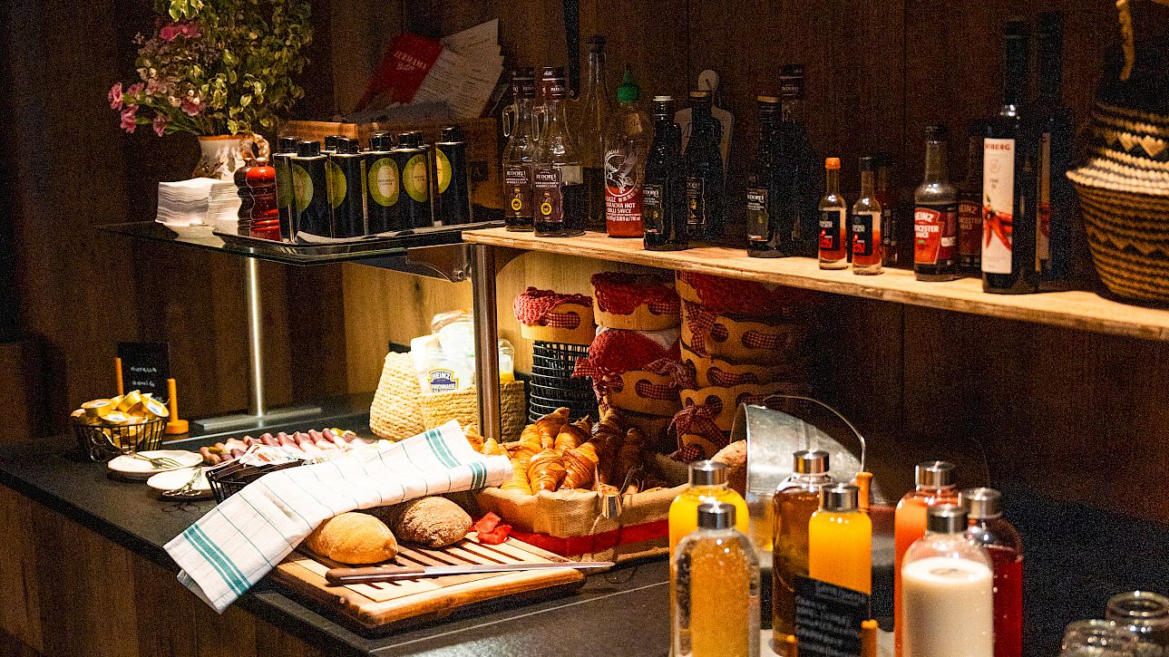 Self-service buffet with juice, pastries, and bread in a rustic setting. Shelves with bottles in the background.