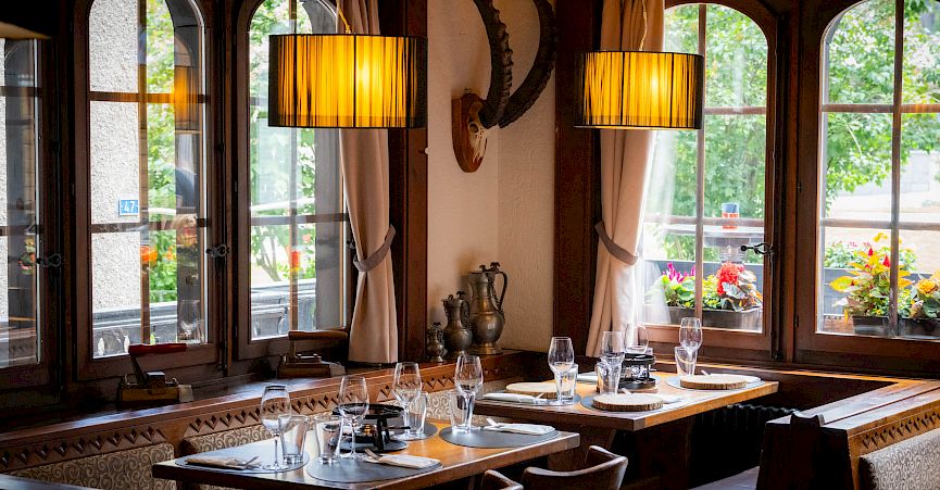 Cozy restaurant with wooden tables set for dining. Large windows overlook blooming plants.