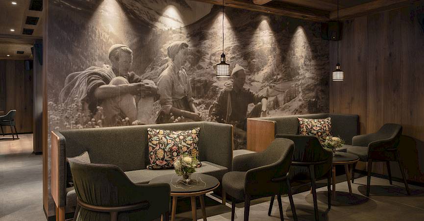 Salon confortable avec canapé et chaises, grande fresque en noir et blanc de travailleurs de montagne. Éclairage tamisé.