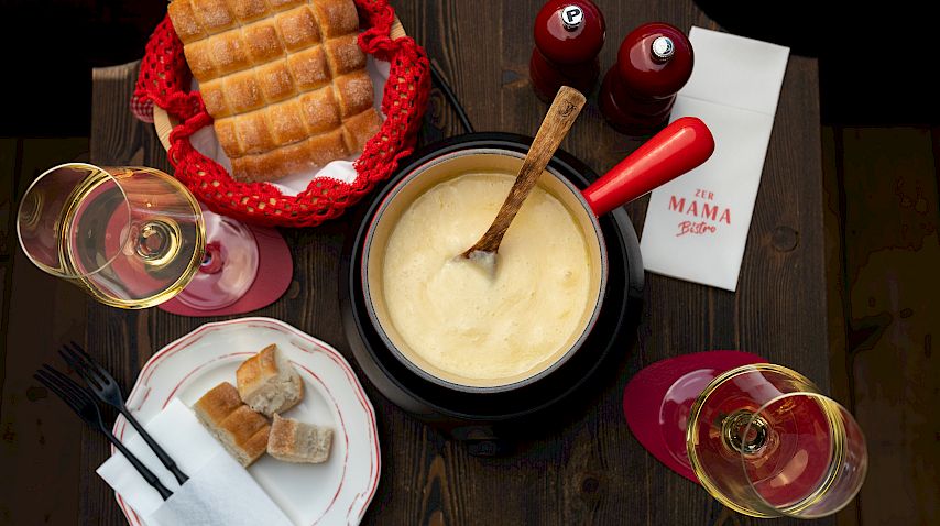 Ein Tisch mit Käsefondue, Brot in einem Korb, zwei Gläsern Wein und zwei roten Salz- und Pfefferstreuern.