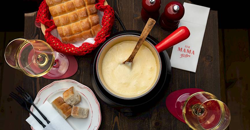 Fondue-Set auf Holztisch mit Brot, zwei Weingläsern und zwei rot-weißen Servietten.