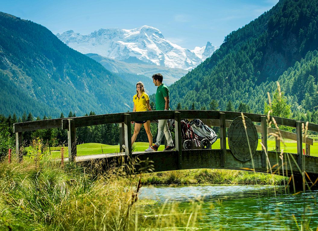 A couple walks over a wooden bridge on a golf course in a mountainous landscape.